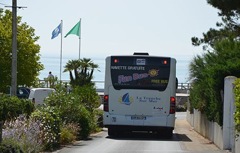 Une navette blanche de La Tranche-sur-Mer s'éloigne sur une rue ensoleillée près de la côte, passant devant des drapeaux et une végétation luxuriante.