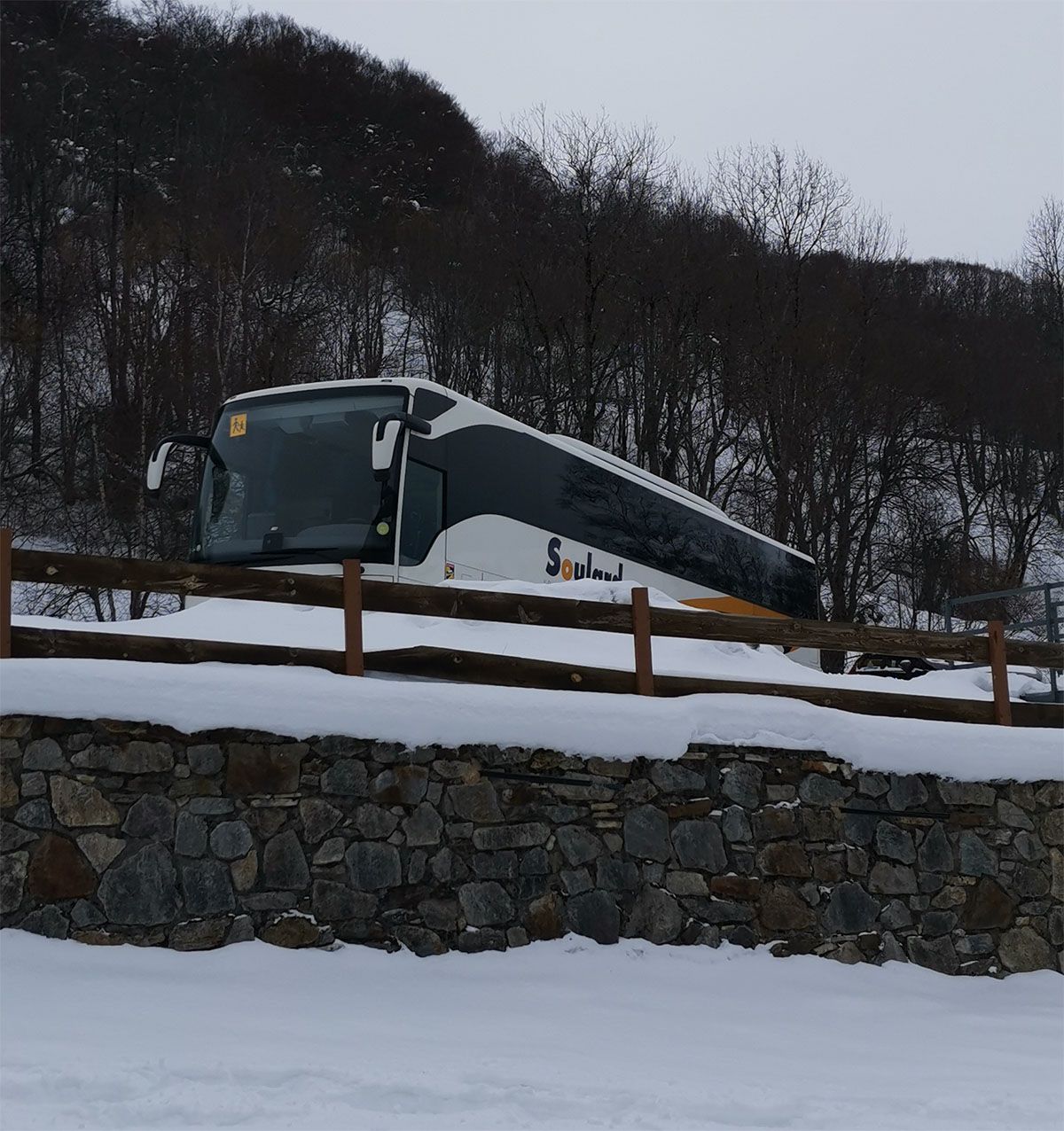 Un autocar blanc et gris garé derrière une clôture en bois, se détachant sur un paysage enneigé et un flanc de colline boisé.