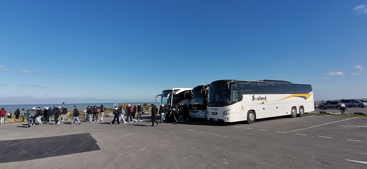 Un groupe de personnes se rassemble près de bus stationnés sur un parking goudronné surplombant l'océan, sous un ciel bleu azur.