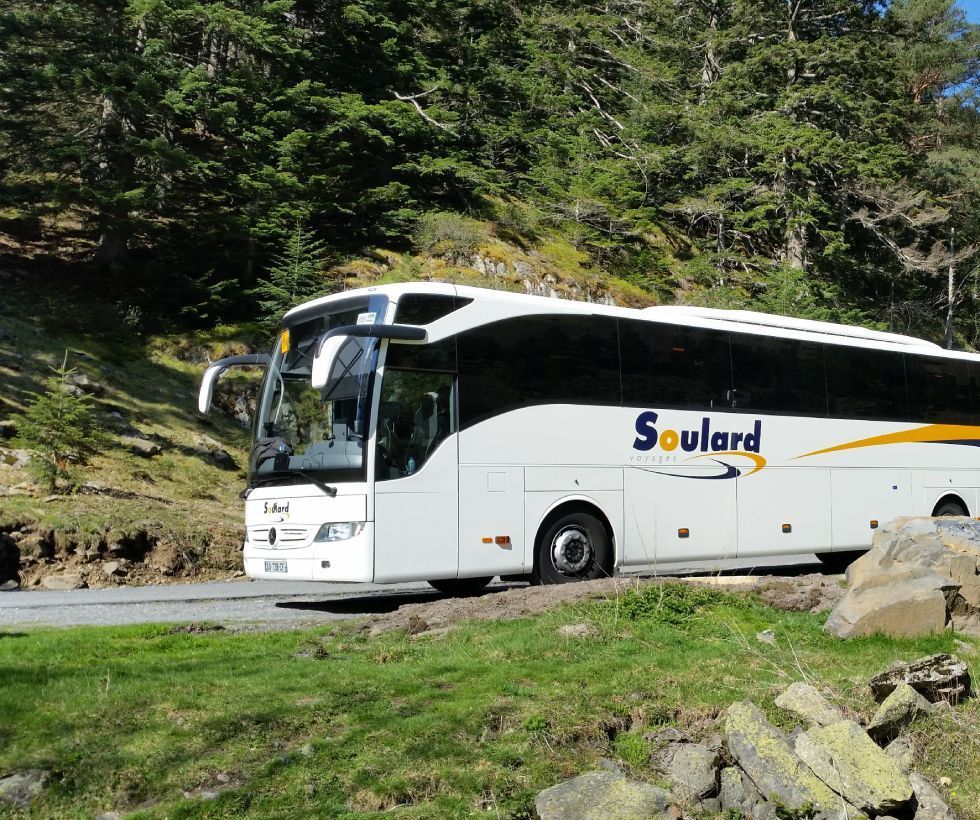 Un bus touristique blanc de la compagnie Soulard est garé sur une route goudronnée, à flanc de colline boisée.