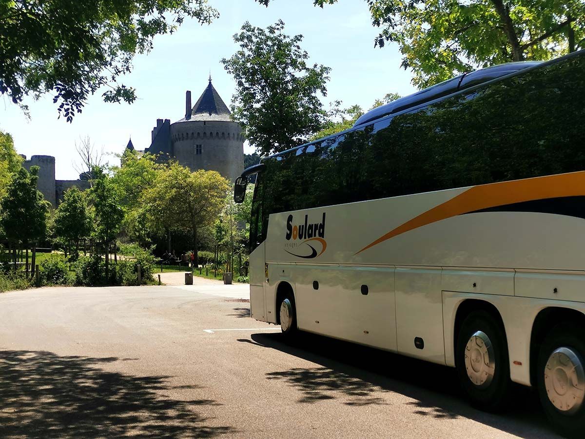 Un bus touristique blanc portant l'inscription « Soule » est garé sur un chemin de gravier à côté d'un château en pierre historique et d'une végétation luxuriante.
