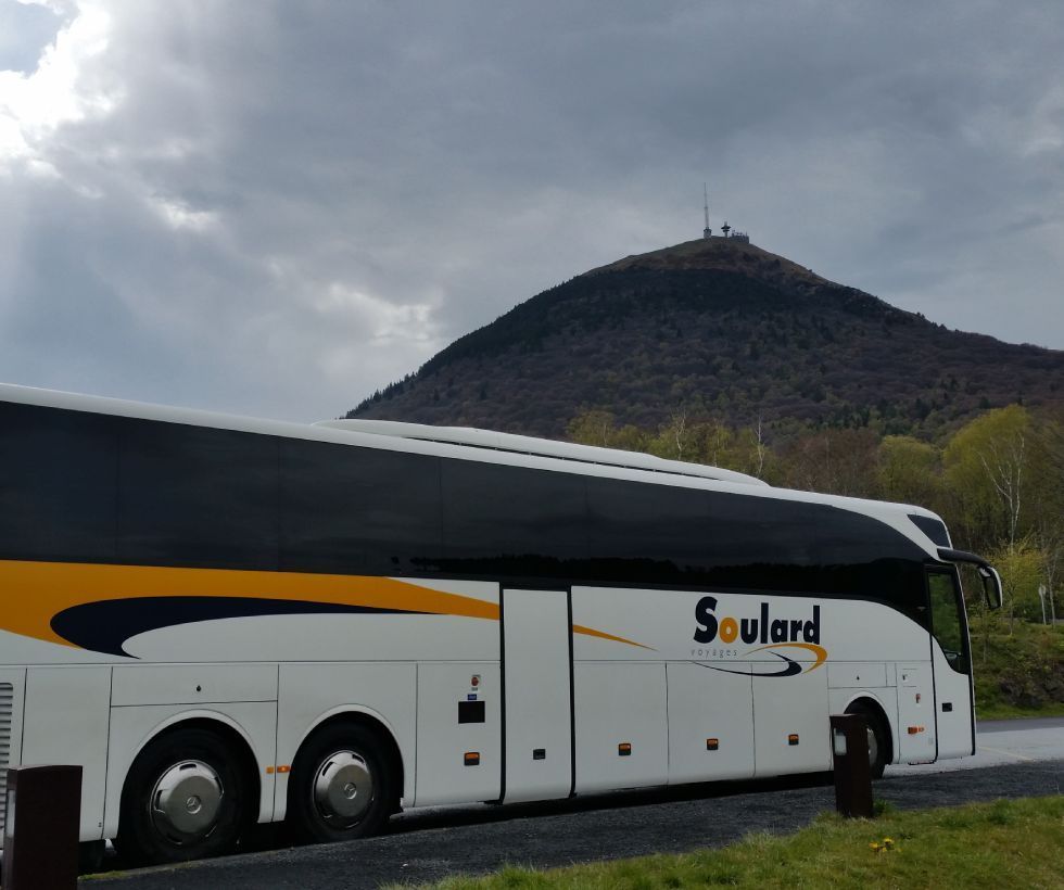 Un autocar blanc à bordures jaunes et noires, portant l'inscription « Soulard », était stationné devant une grande colline surmontée d'une tour de télécommunications.