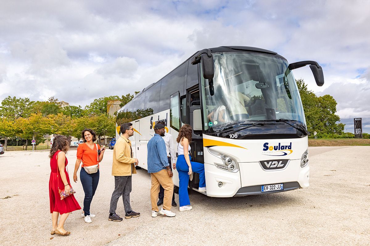 Quatre personnes montent à bord d'un bus touristique blanc portant la marque « Soule » sur un parking en gravier.
