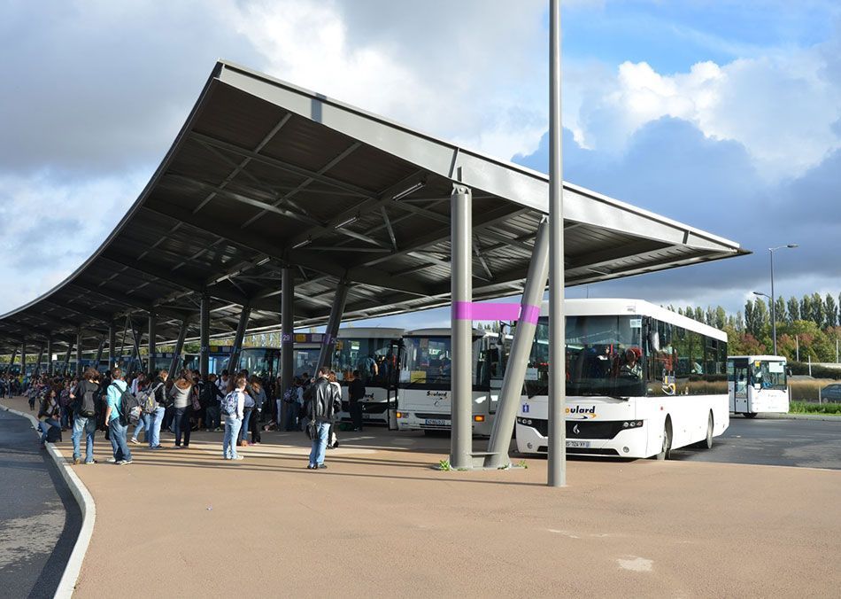 Des personnes patientent dans une gare routière couverte, sous un grand auvent métallique incurvé, où plusieurs bus blancs sont stationnés.