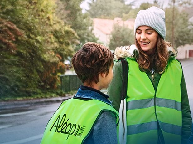 Deux personnes portant des gilets de sécurité vert fluo se tiennent à l'extérieur, près d'une route, et semblent discuter entre elles.