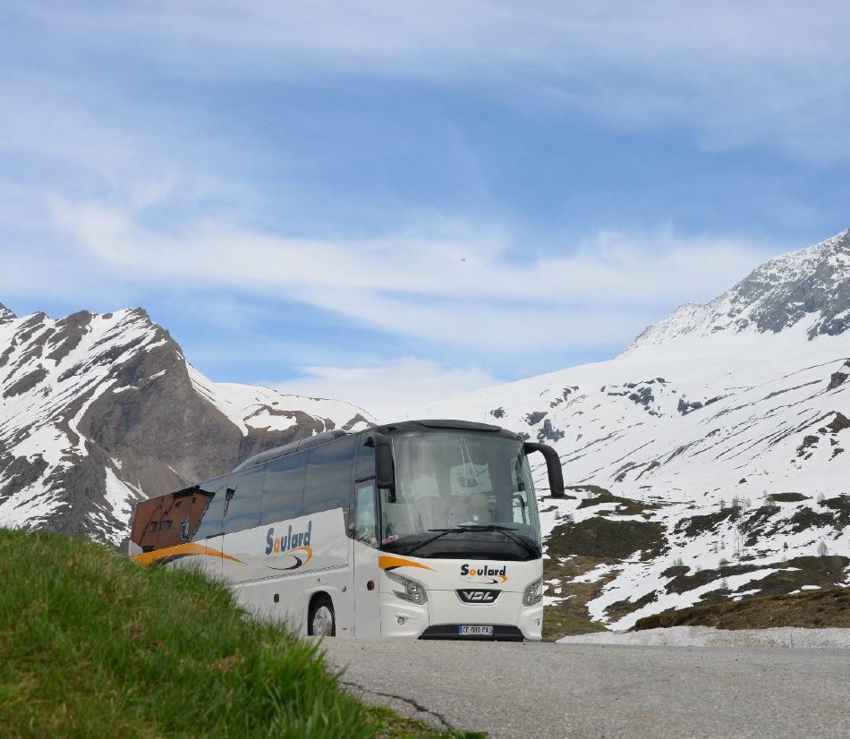 Un bus touristique blanc roule sur une route de montagne asphaltée, avec des sommets enneigés en arrière-plan.