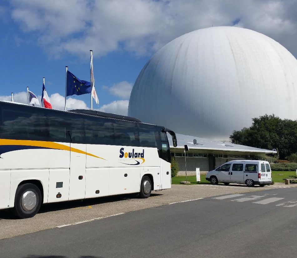 Un bus Soulard garé devant un grand bâtiment blanc en forme de dôme, sous un ciel bleu ensoleillé parsemé de drapeaux.