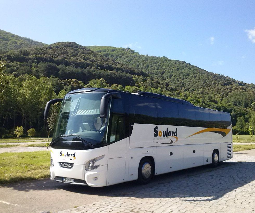 Un bus touristique blanc de la compagnie Soulard est garé sur un parking goudronné, avec une colline boisée et verdoyante en arrière-plan, sous un ciel bleu.