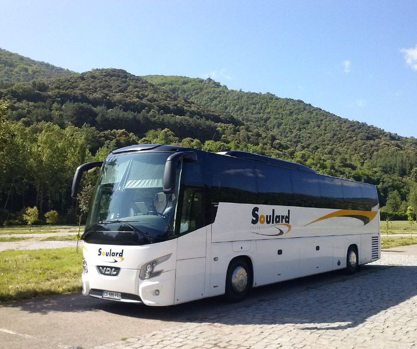 Un bus touristique blanc de la compagnie Soulard est garé sur un parking goudronné, avec une colline boisée et verdoyante en arrière-plan, sous un ciel bleu.