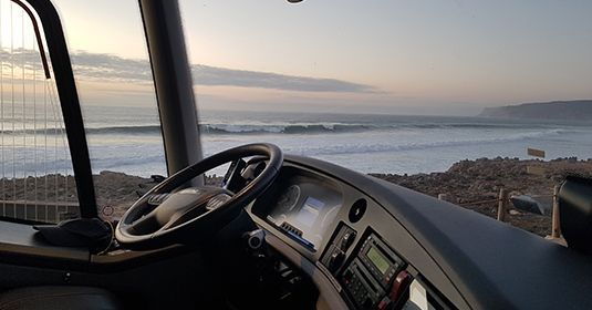 Vue depuis la cabine d'un camion, donnant sur une plage aux vagues déferlantes sous un ciel pâle au coucher du soleil.