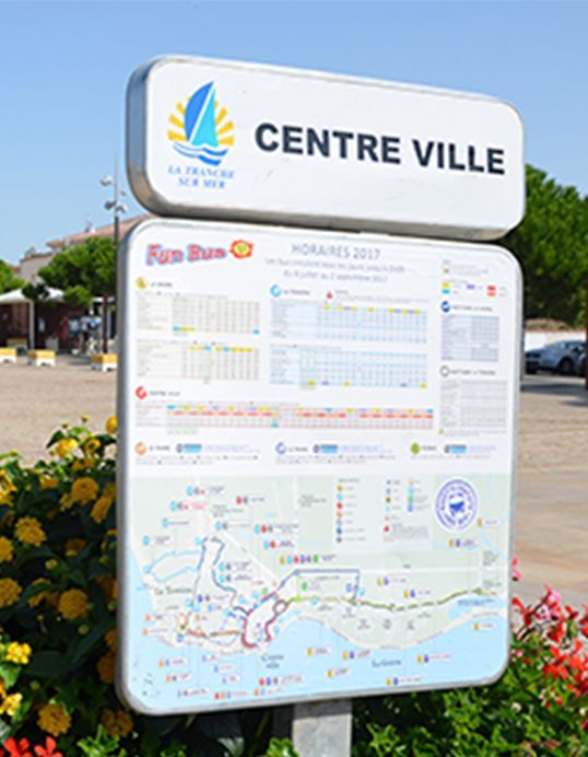 Un panneau d'information en centre-ville avec un plan des transports en commun et les horaires de bus de 2017 à La Tranche-sur-Mer, installé à l'extérieur au milieu de la végétation.