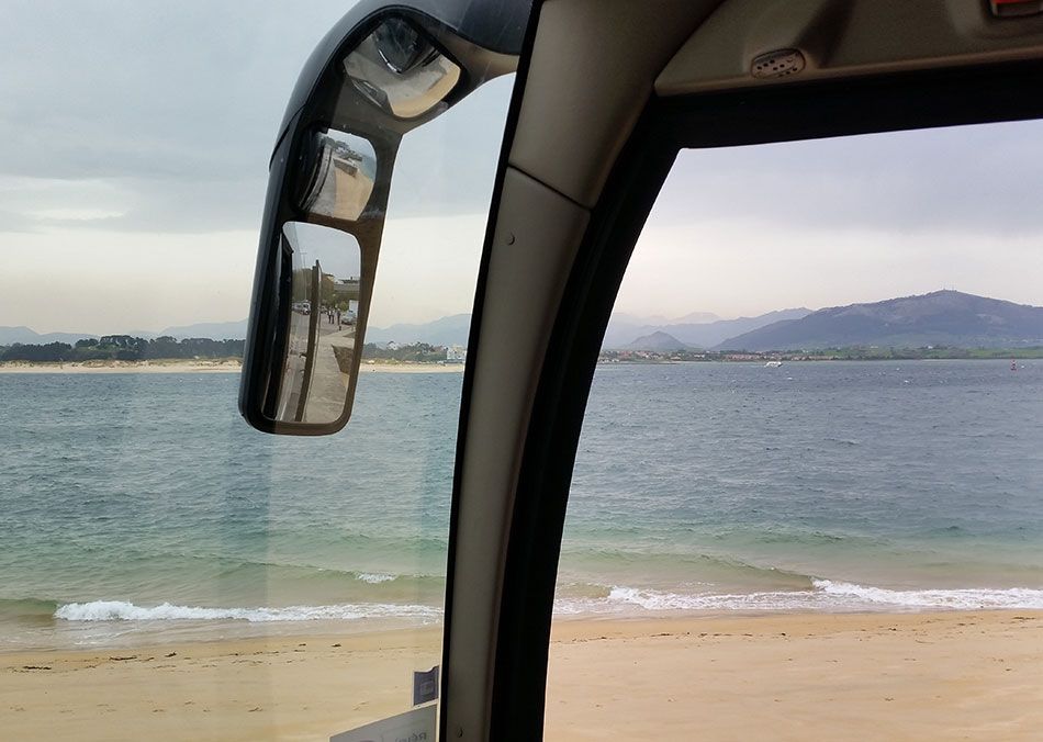 Vue depuis la fenêtre d'un bus montrant une plage de sable fin, une eau calme et des montagnes au loin sous un ciel nuageux.