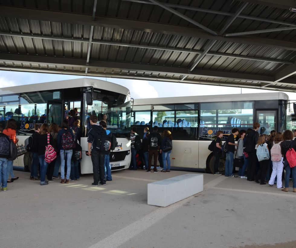 En journée, un groupe de personnes monte à bord de deux bus touristiques blancs stationnés sous un terminal abrité.