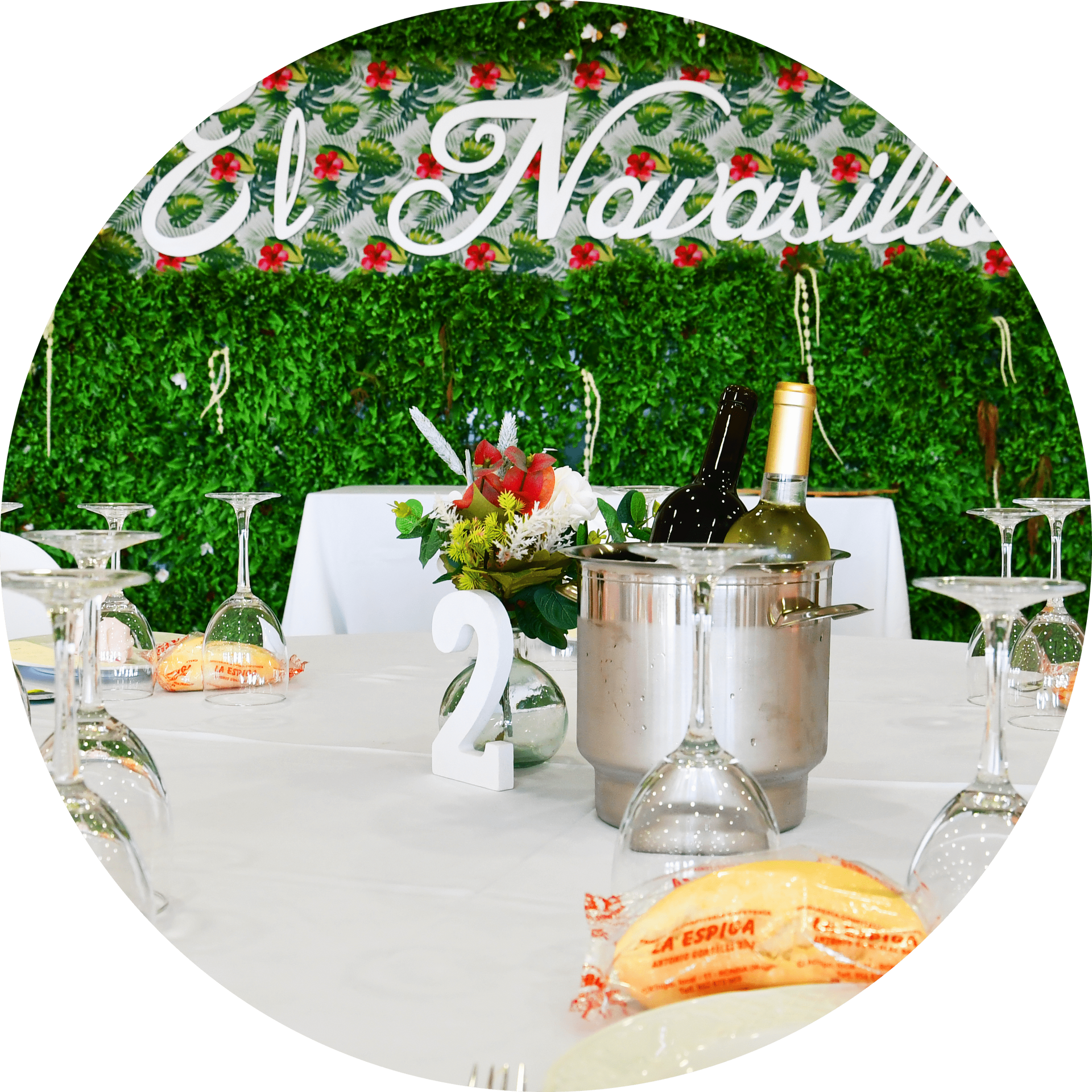 Montaje de mesa para celebración, con vino, pan y centros florales bajo un cartel que dice