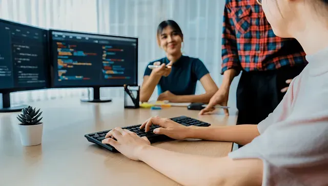 Una mujer está escribiendo en un teclado frente a dos monitores de computadora.