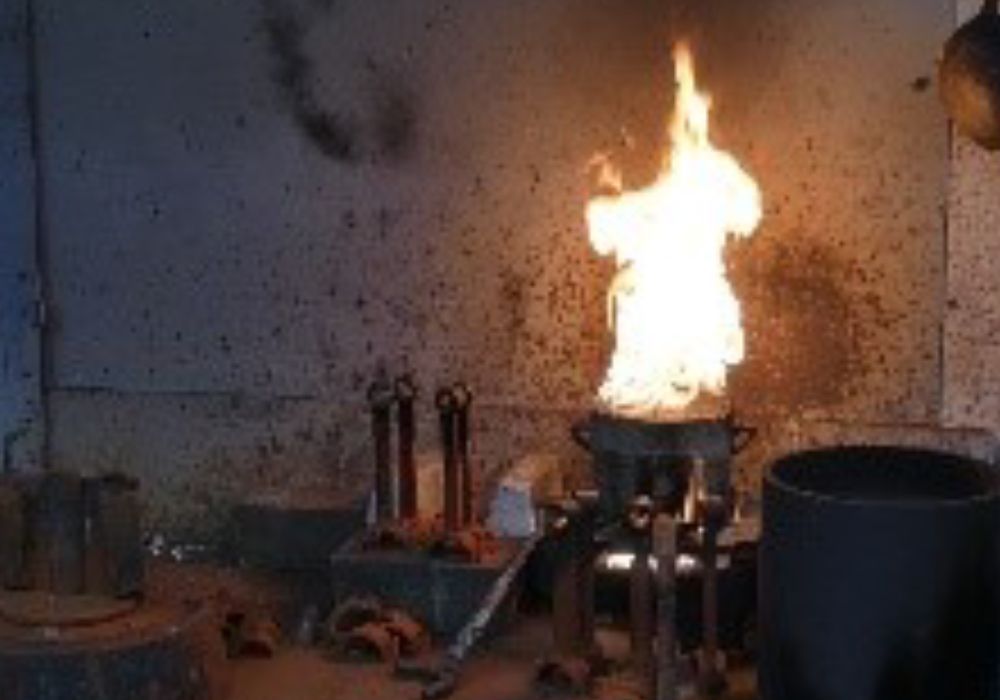 Une flamme brûle dans un récipient métallique, dans un espace industriel faiblement éclairé, avec des outils à proximité.