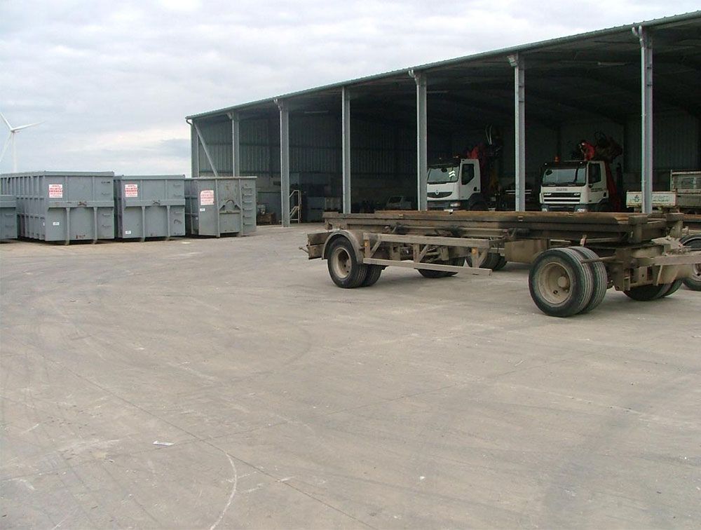 Bennes grises, châssis de camions et camions bâchés sous un hangar sur un terrain en béton. Ciel couvert.