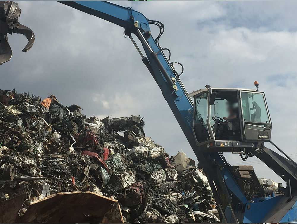 Une grue à griffes bleues soulève des déchets métalliques d'un grand tas dans une casse.