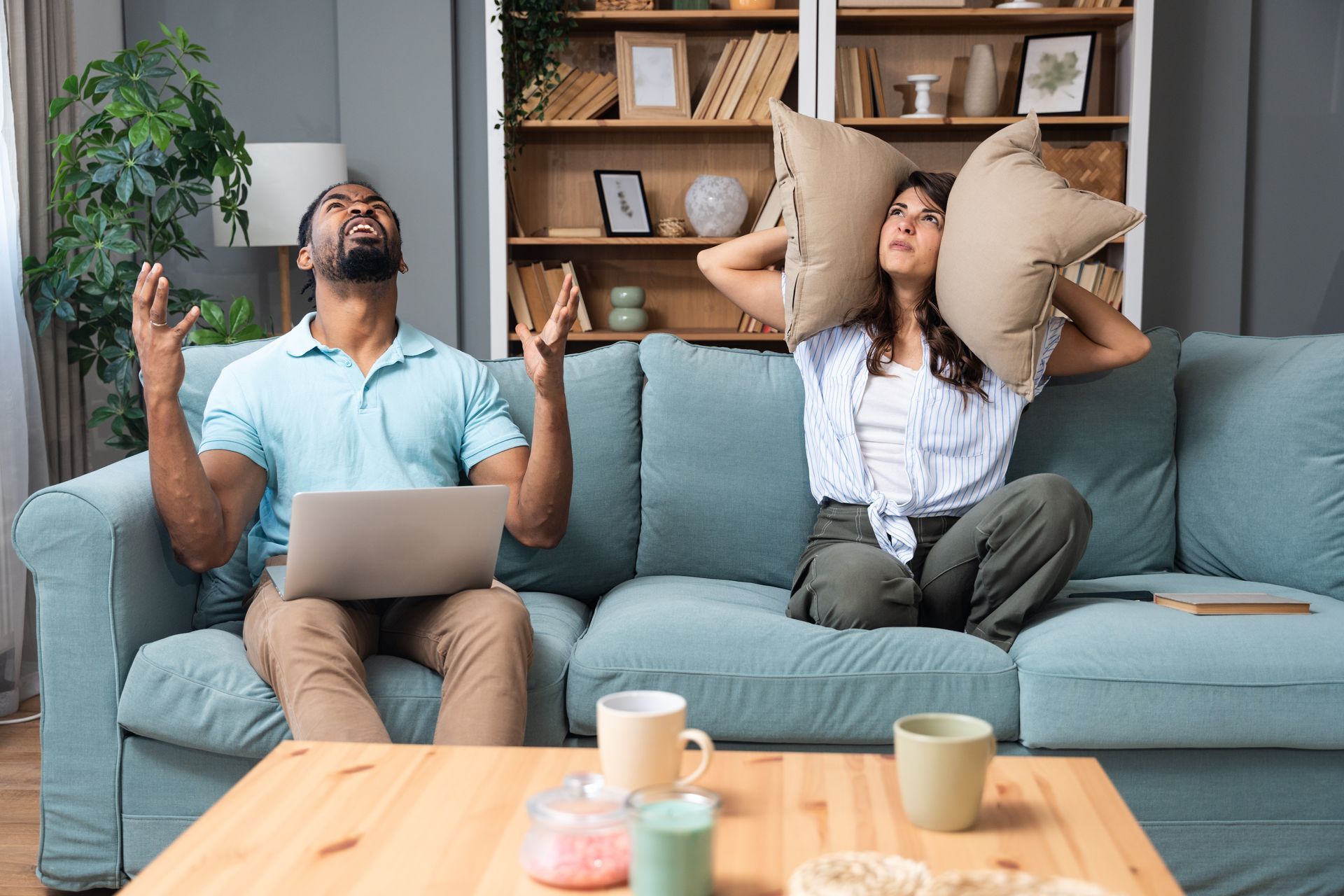 Un homme travaille sur son ordinateur portable ; une femme, assise sur le canapé, se bouche les oreilles avec des oreillers.