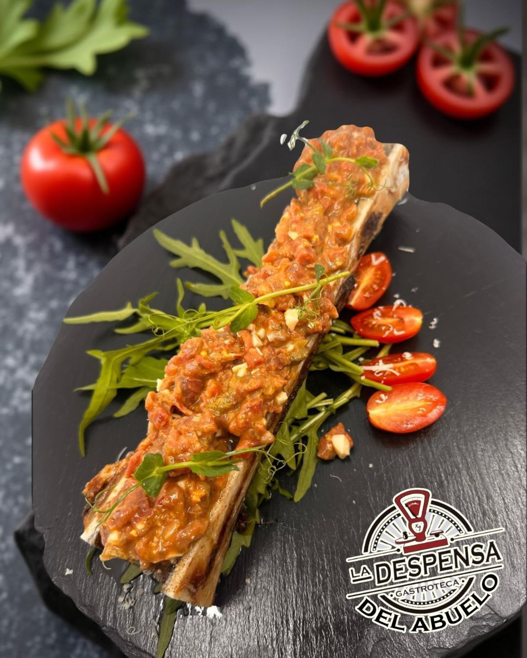 Médula ósea asada cubierta con salsa de tomate, rúcula y tomates cherry en un plato de pizarra.
