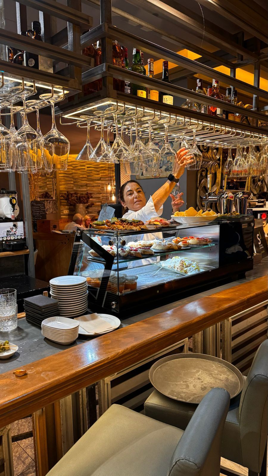 Mujer detrás de una barra buscando vasos, exhibición de comida, iluminación cálida y acentos de madera.