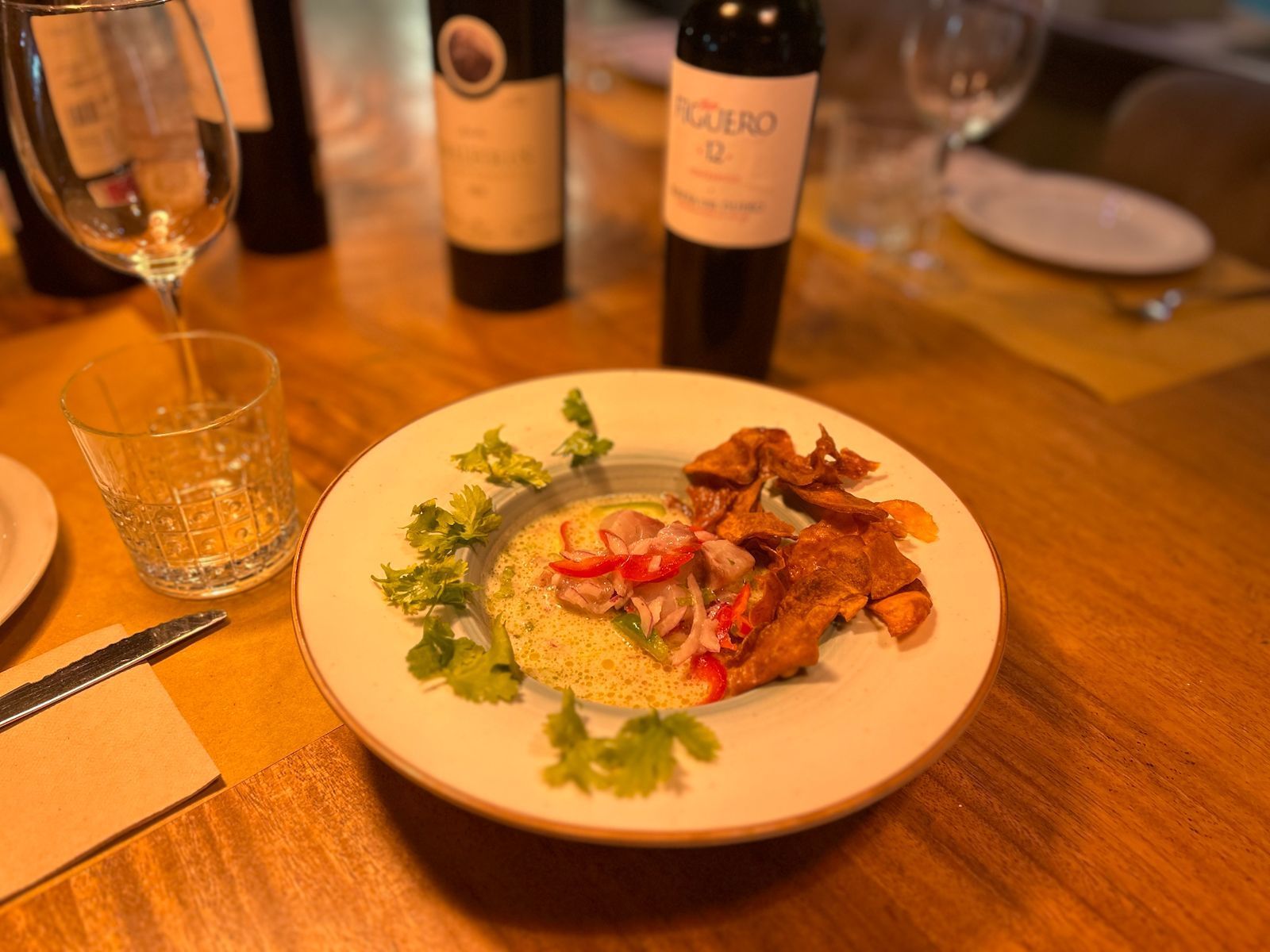 Un plato de comida con elementos rojos y beige sobre una mesa con copas de vino y botellas al fondo.