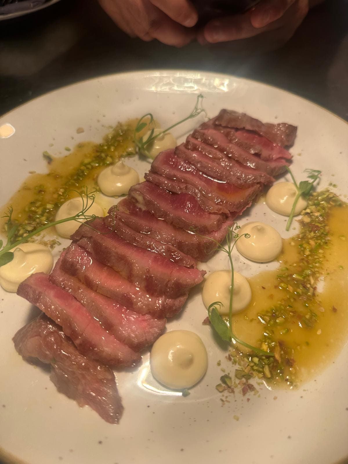 Filete cortado en rodajas en un plato blanco, adornado con salsa, pequeños bulbos blancos y hierbas verdes.