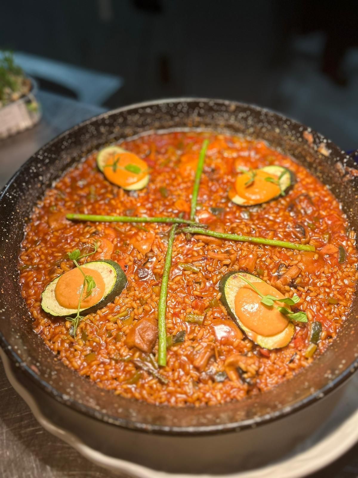 Paella en sartén negra, cubierta con rodajas de calabacín y salsa de naranja, adornada con cebollino.