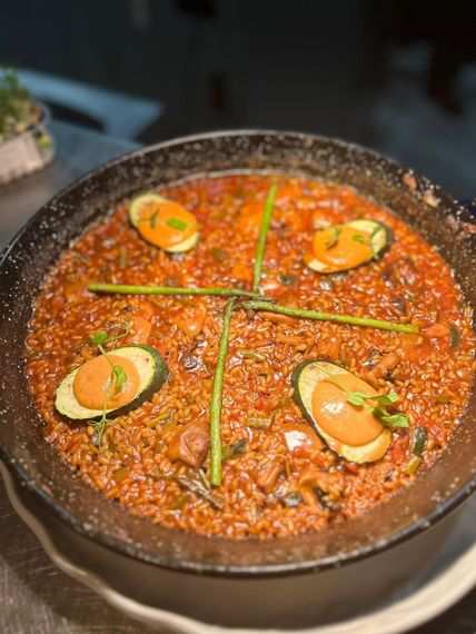 Paella en sartén negra, cubierta con rodajas de calabacín y salsa de naranja, adornada con cebollino.