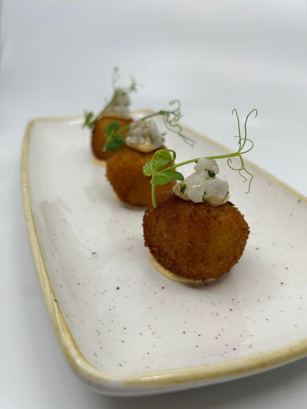 Tres croquetas de color marrón dorado cubiertas con salsa blanca y brotes de guisantes en un plato rectangular.