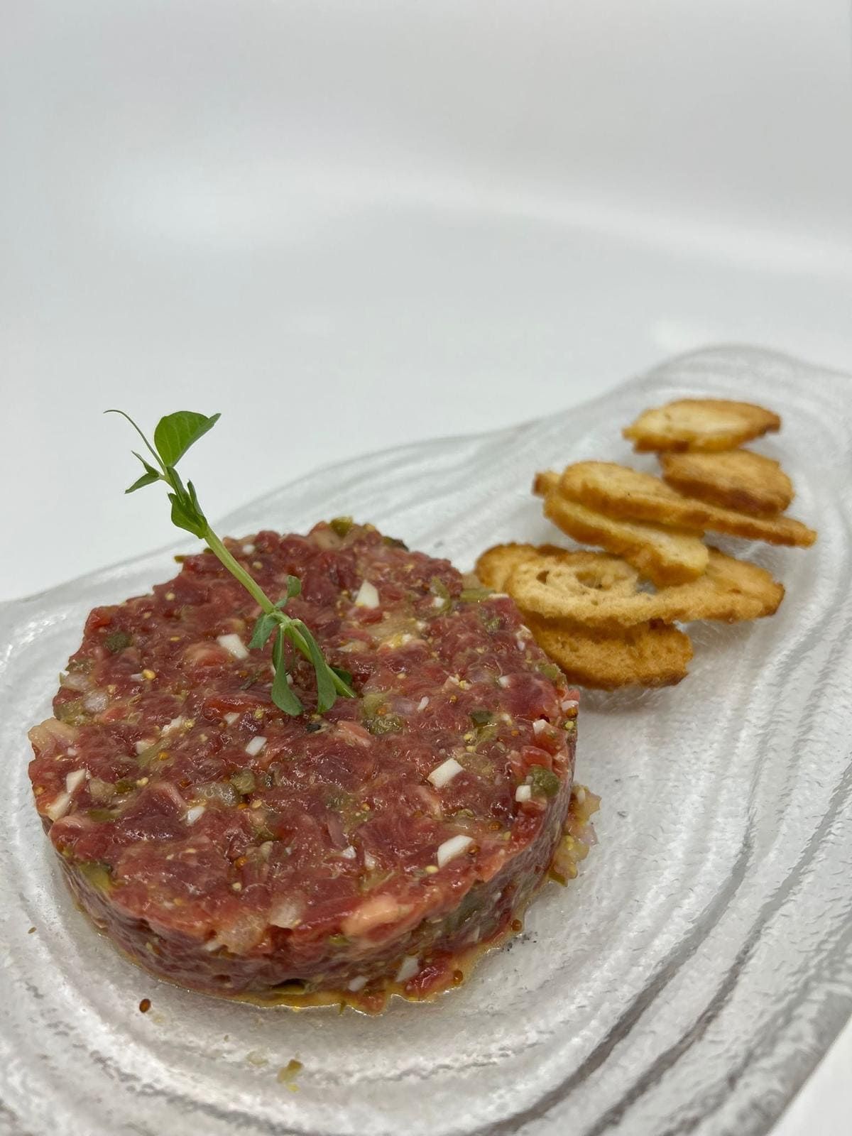 Tártaro de ternera con picatostes, adornado con una ramita de hierbas verdes, en un plato transparente y ondulado.