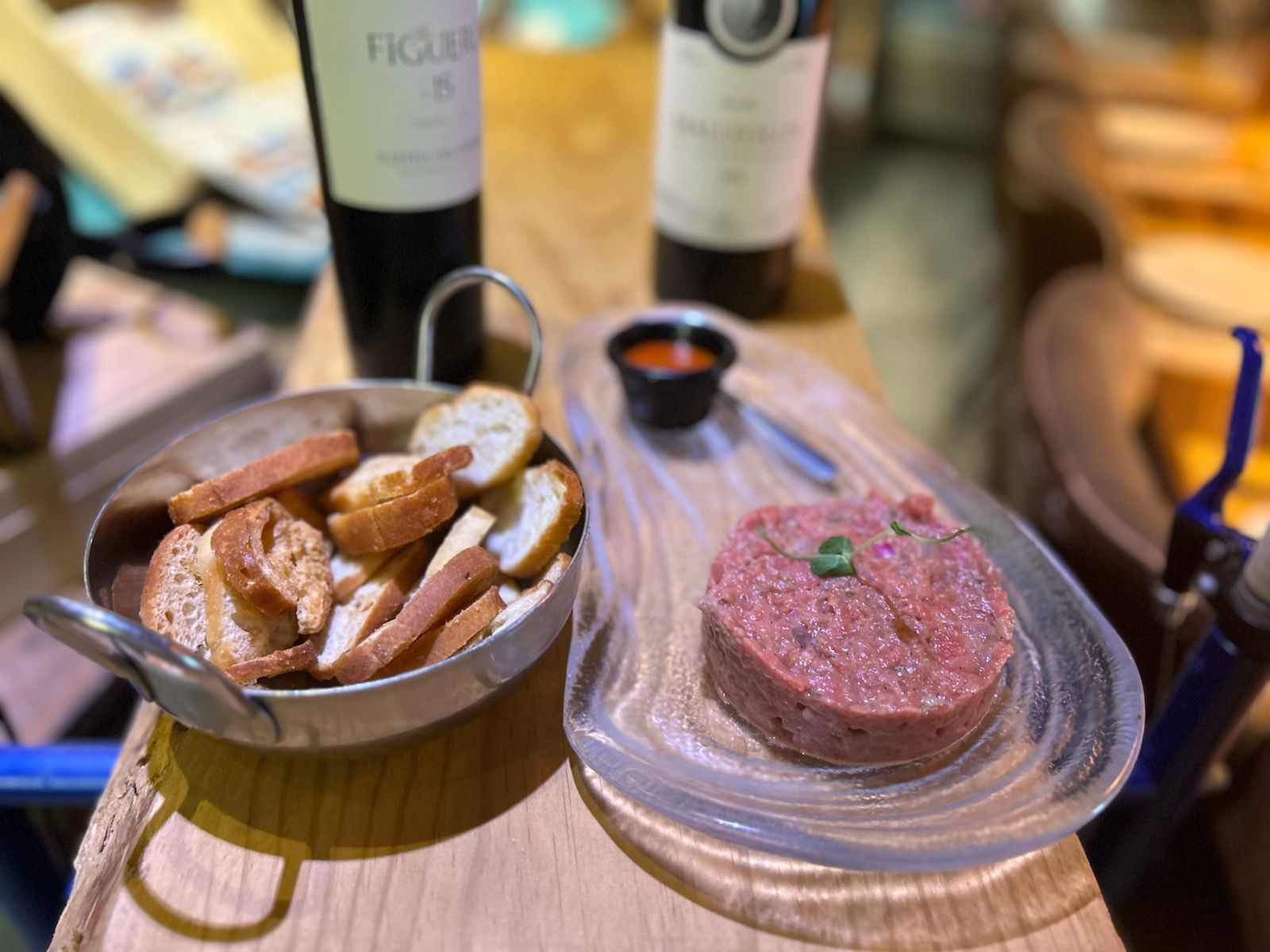 Steak tartar con puntas tostadas, salsa roja, botellas de vino sobre una mesa de madera.