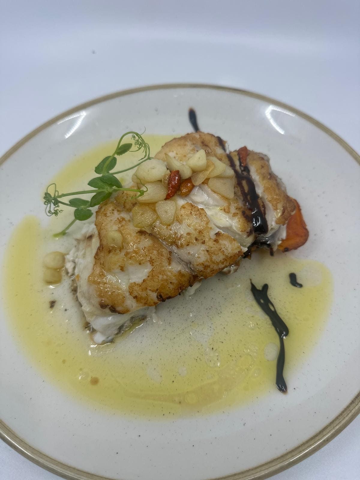 Pescado sellado con costra dorada, servido en un plato con una salsa ligera y guarnición de brotes.