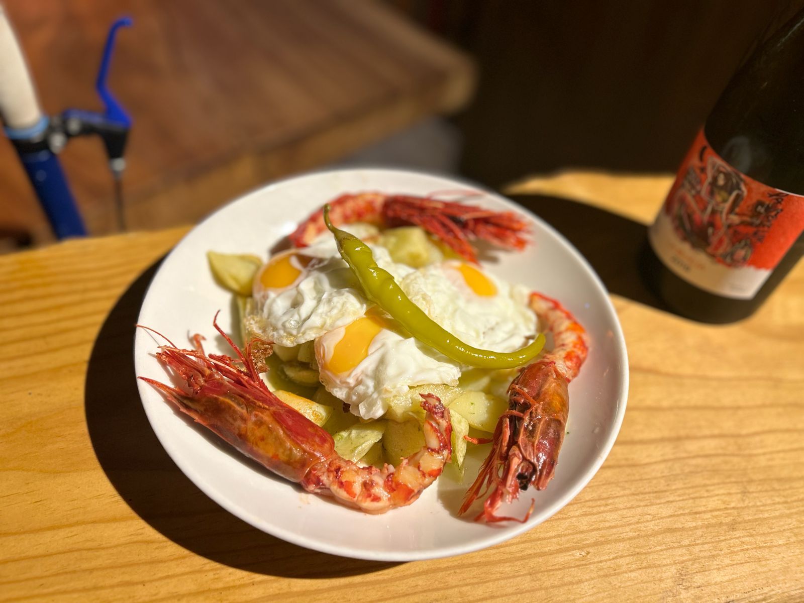 Plato de huevos fritos, camarones, patatas y pimientos con una botella de vino sobre una mesa de madera.