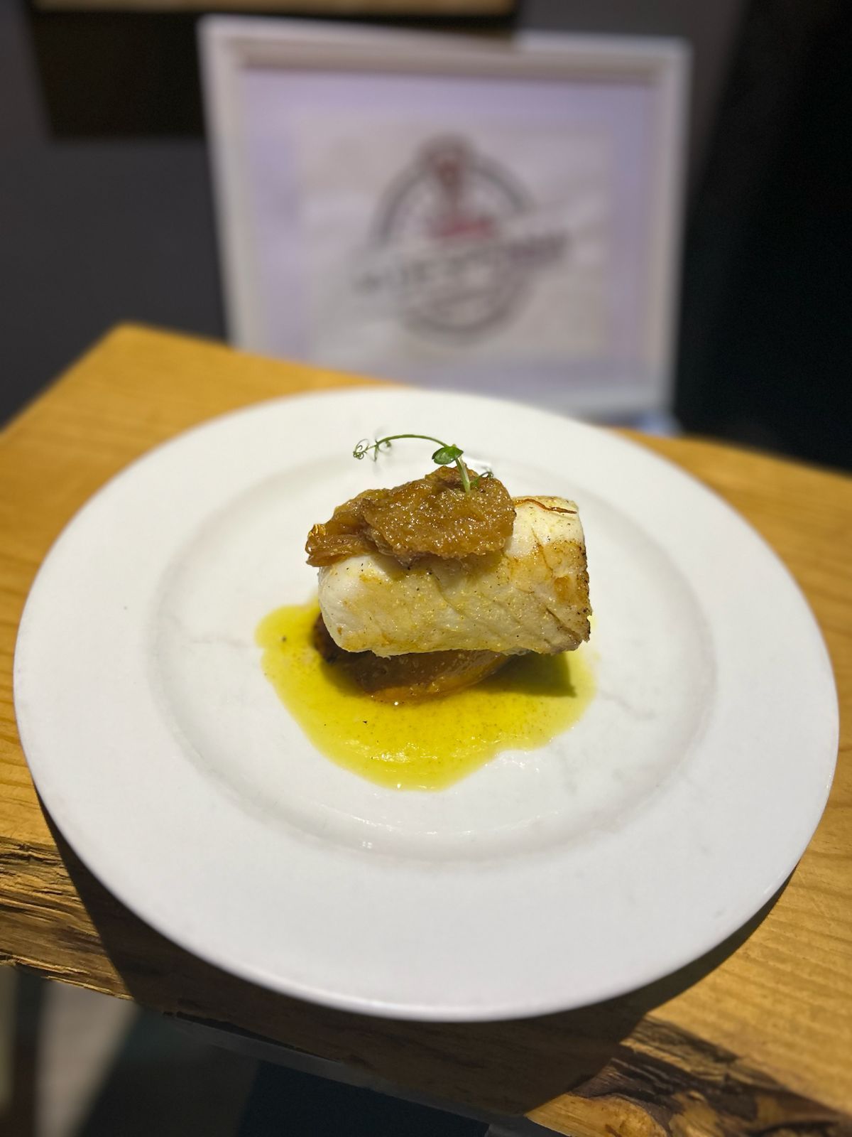 Plato blanco con pescado sellado, salsa y guarnición sobre una mesa de madera.