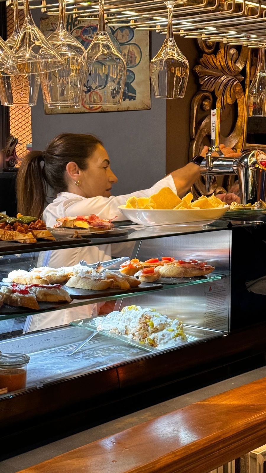 Mujer tras la barra, colocando comida en una vitrina. Vasos colgados encima.