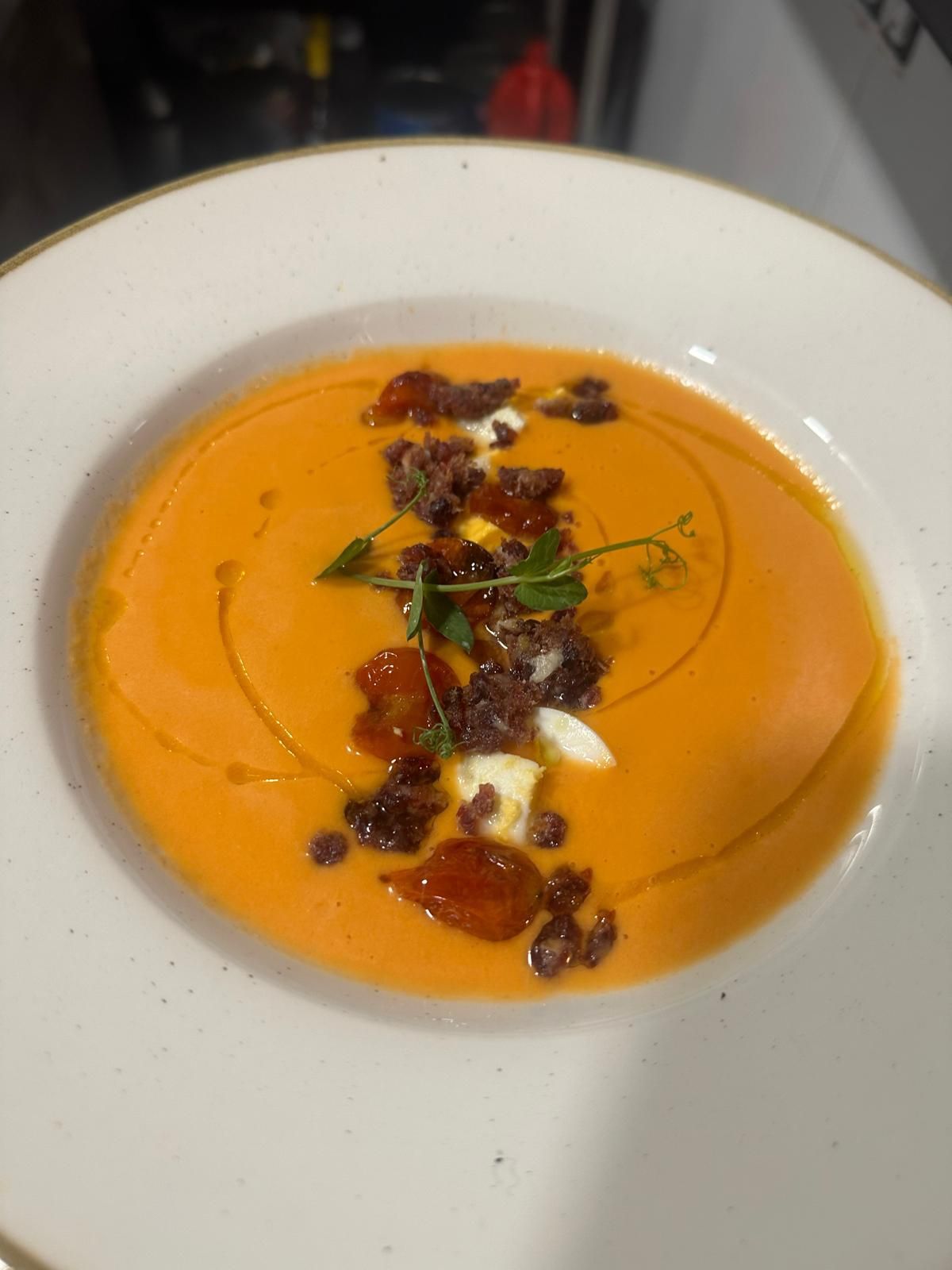 Sopa de naranja en un tazón blanco, adornada con crumble marrón, crema blanca, un chorrito de aceite y brotes verdes.