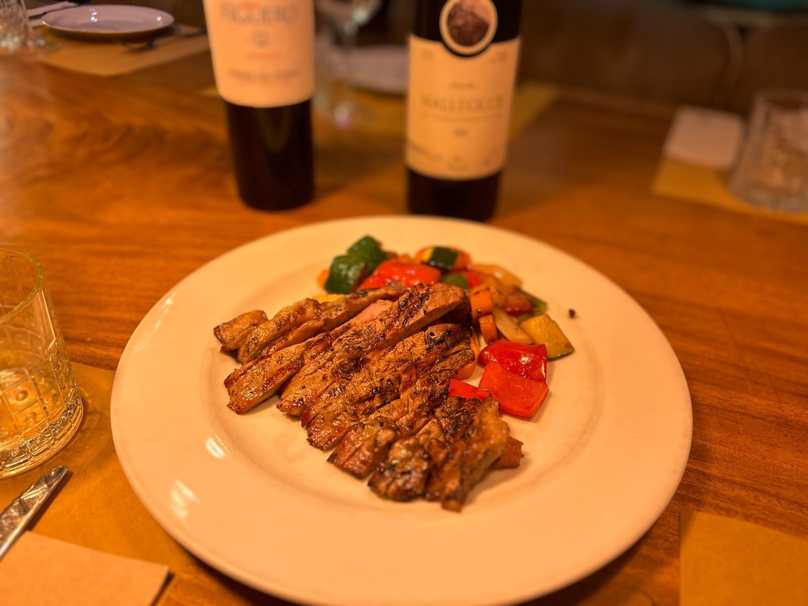 Filete en rodajas con verduras asadas en un plato blanco, con dos botellas de vino de fondo.