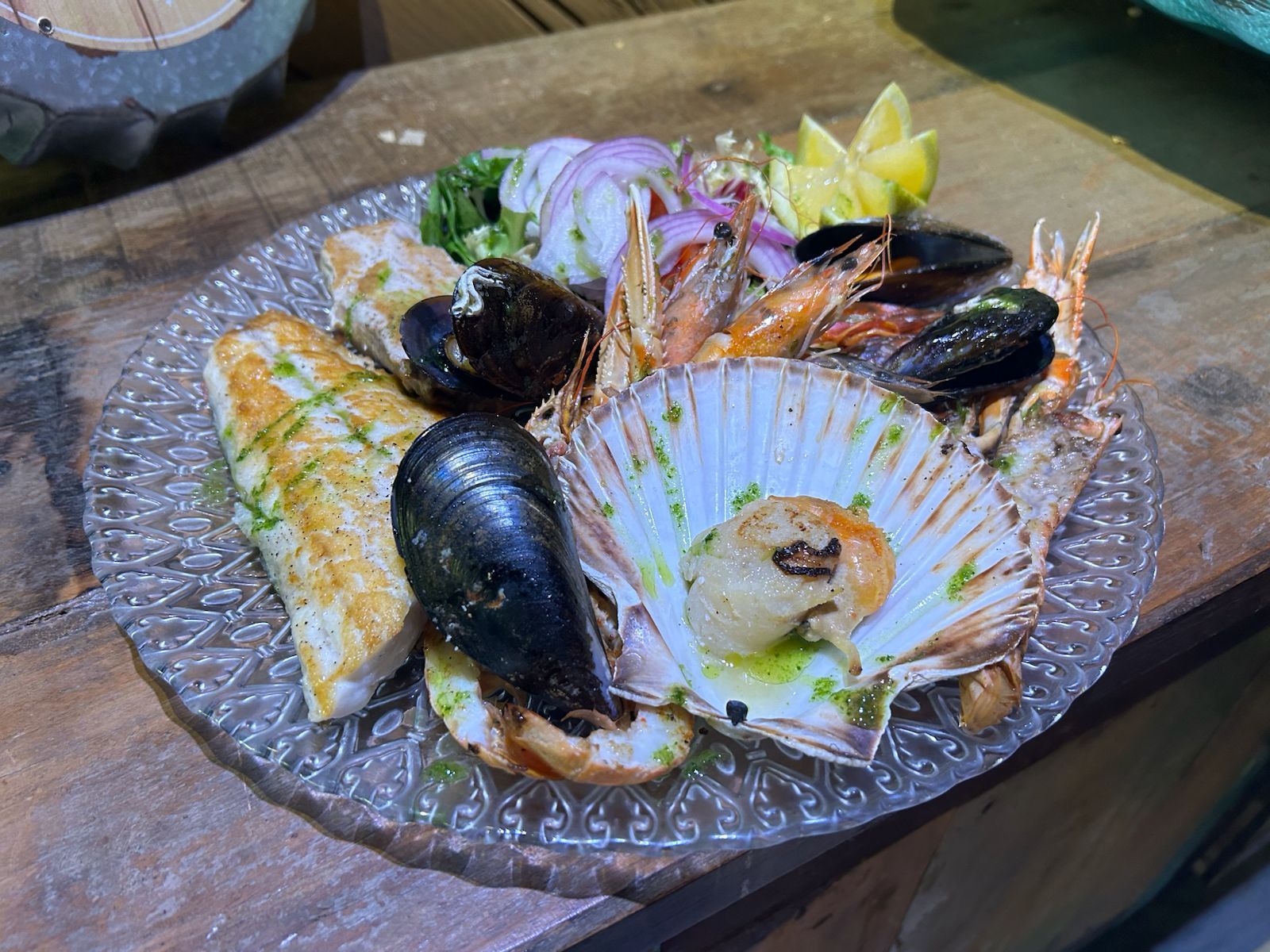 Plato de mariscos: pescado a la parrilla, mejillones, camarones, vieiras, cebollas, limón y hierbas en un plato transparente.