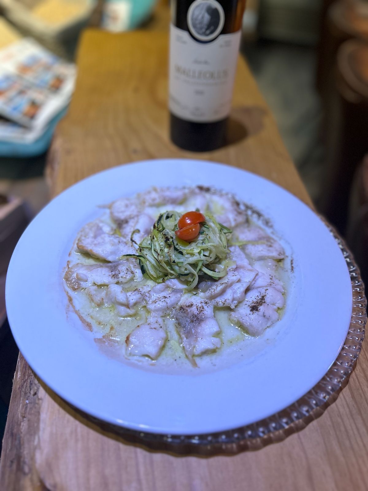 Plato de raviolis con pesto y tomate cherry, botella de vino al fondo.