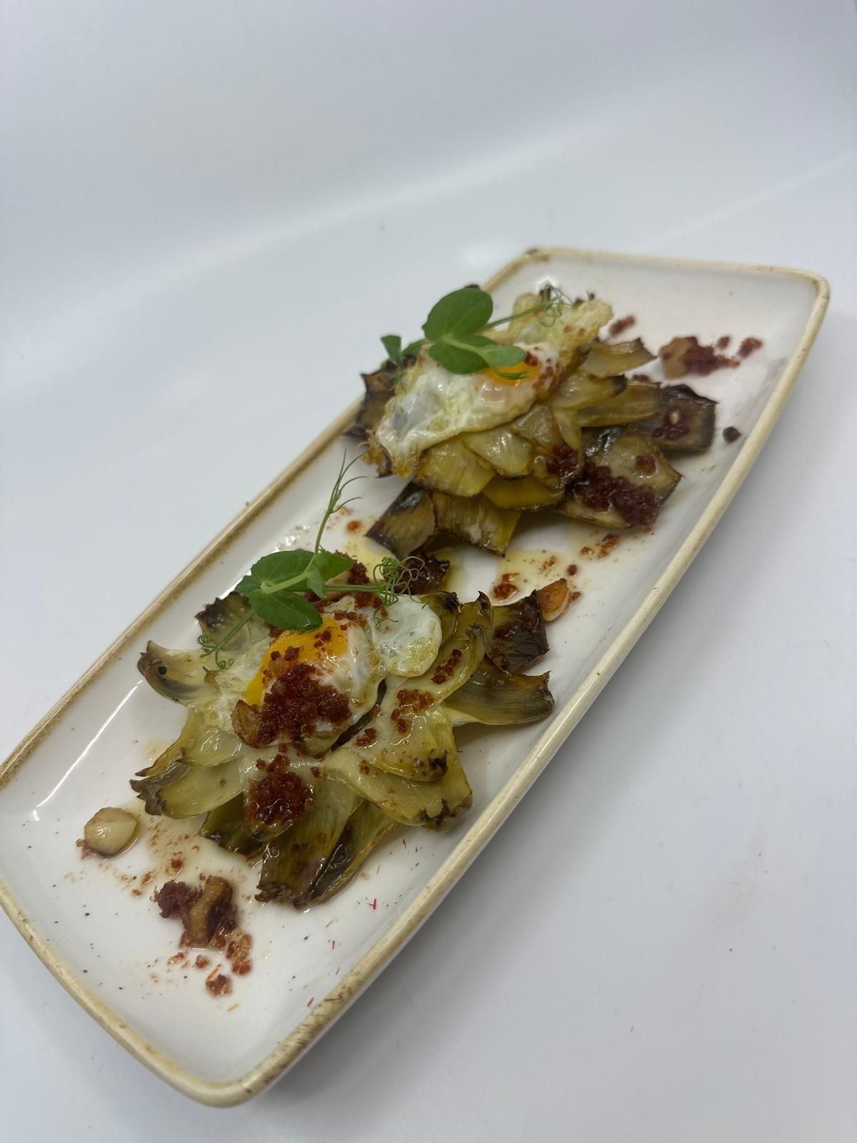 Dos corazones de alcachofa fritos cubiertos con huevos fritos y chorizo en un plato rectangular.