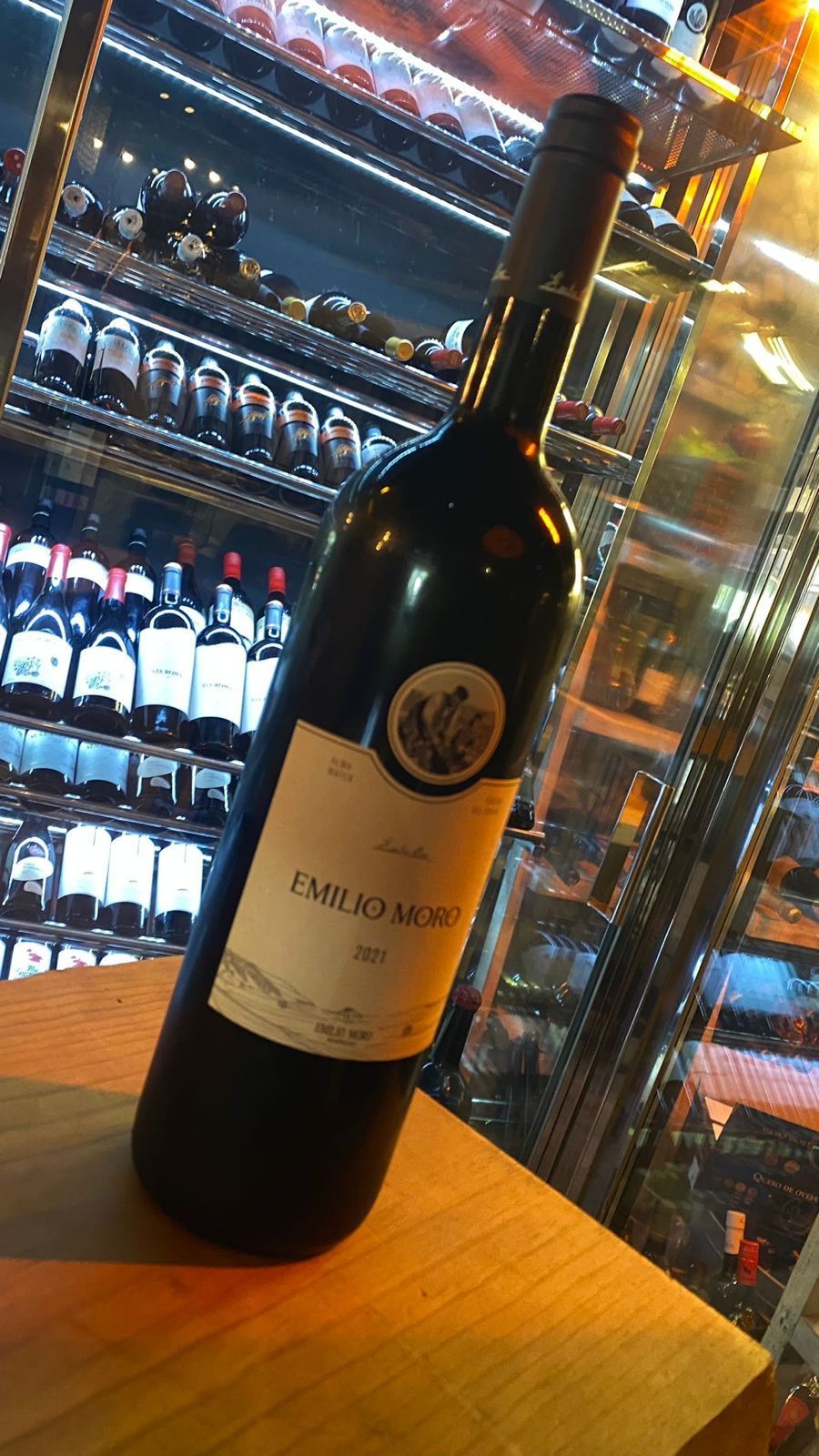 Botella de vino tinto sobre una superficie de madera delante de una vitrina de vinos.