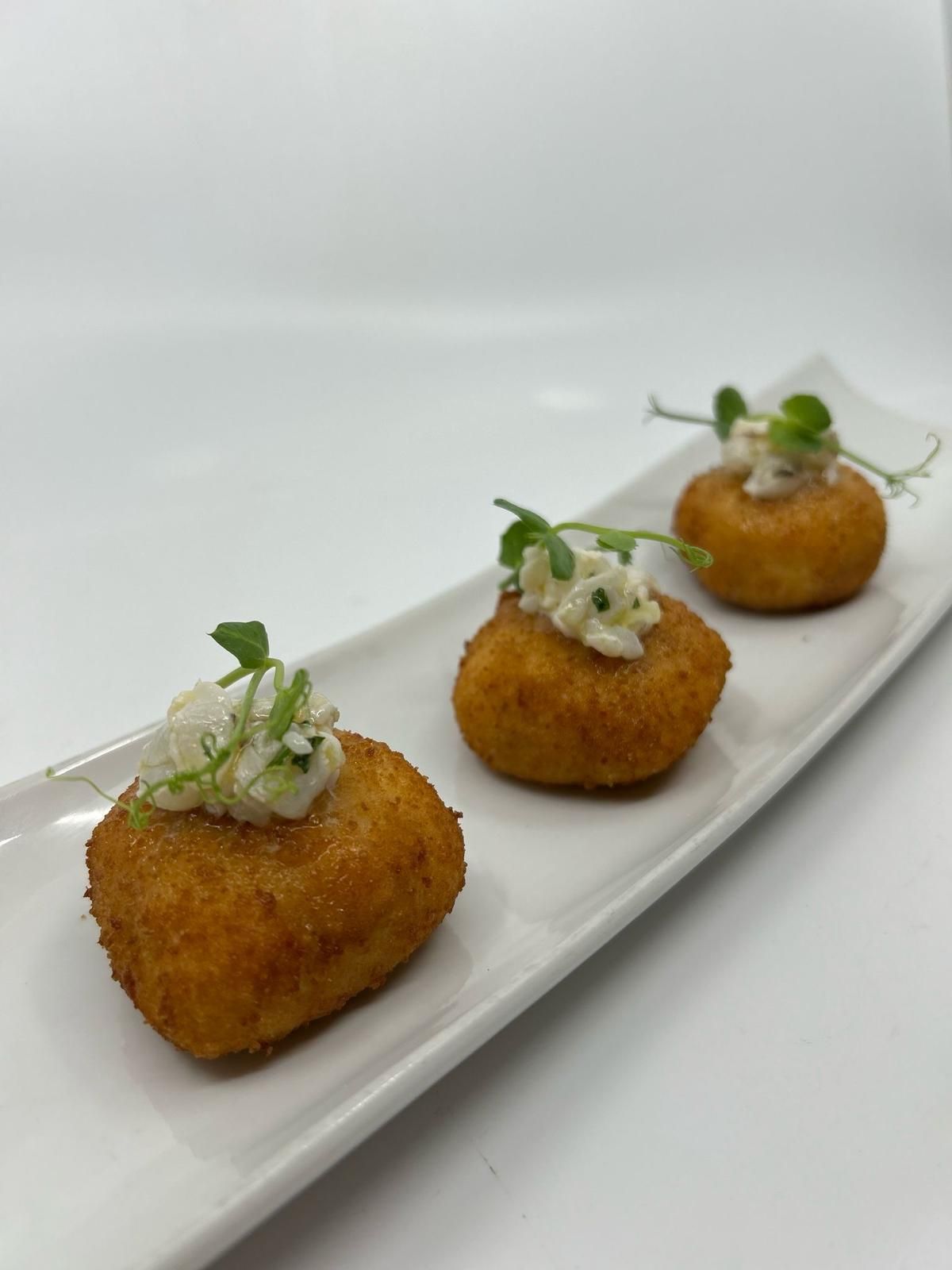Tres aperitivos fritos con cobertura blanca en un plato rectangular, adornados con brotes.