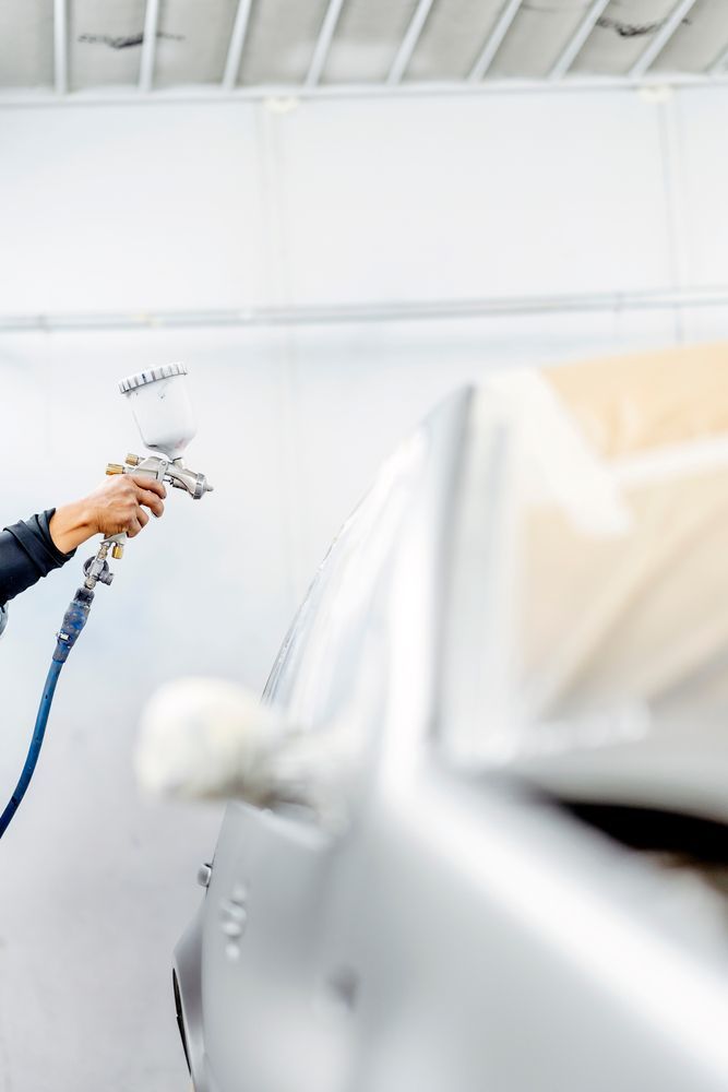 En el interior de un taller de carrocería profesional, una persona utiliza una pistola pulverizadora para aplicar pintura plateada en el lateral de un coche.