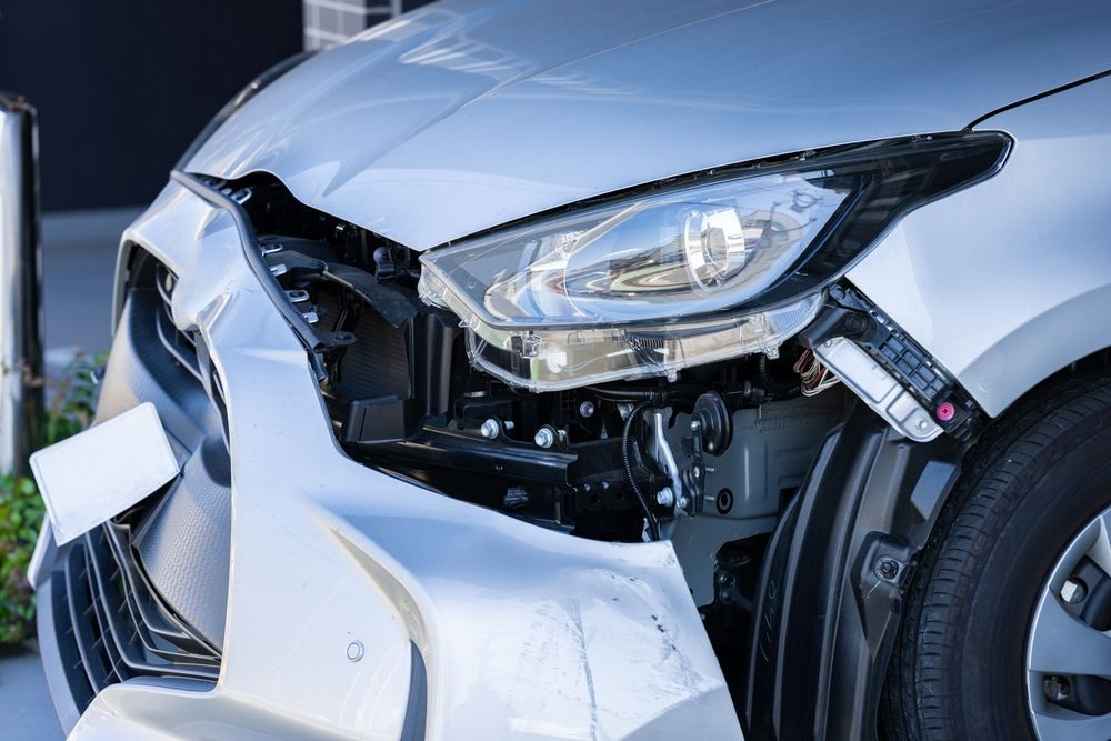 Un coche plateado con importantes daños por colisión en la parte delantera, incluyendo un parachoques desprendido y componentes interiores expuestos.