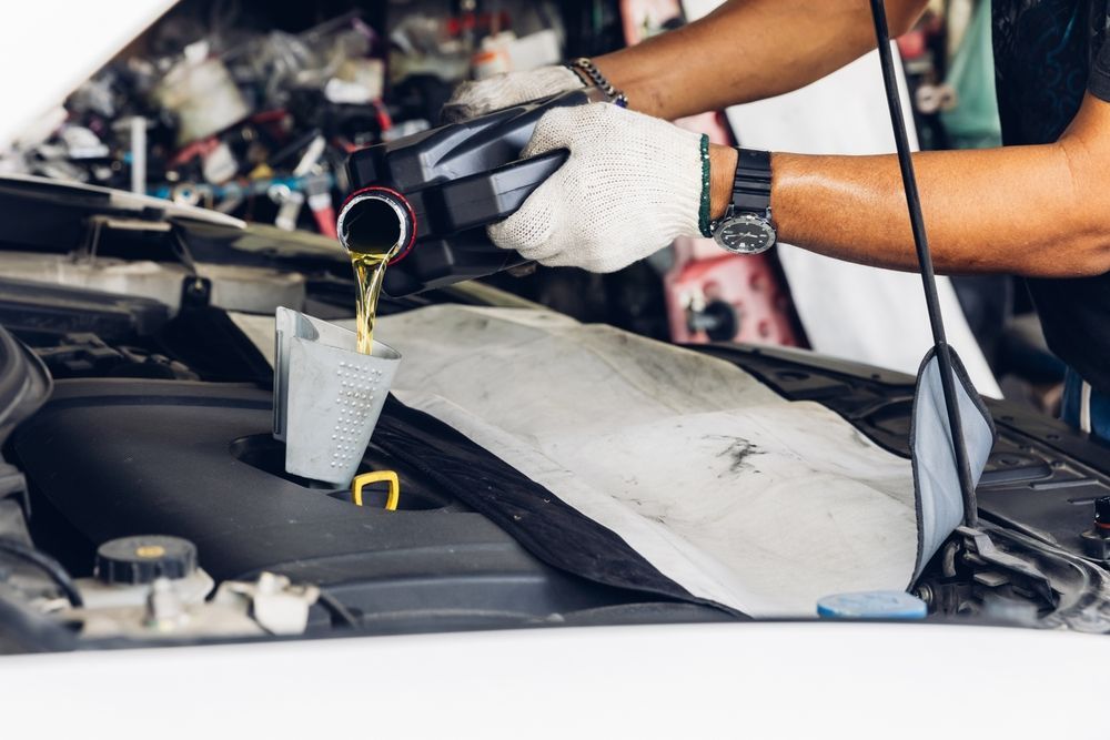 Un mecánico, con guantes de trabajo, vierte aceite de motor nuevo desde un recipiente de plástico negro al motor de un coche utilizando un embudo.