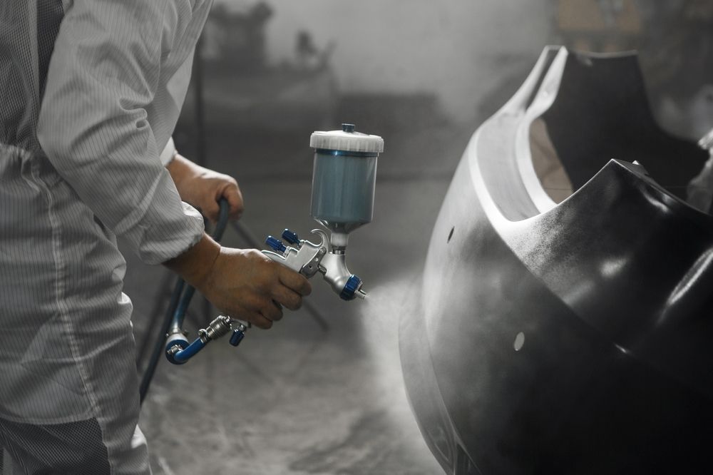 Un trabajador con un traje protector blanco utiliza una pistola pulverizadora para pintar el parachoques oscuro de un coche en un taller.