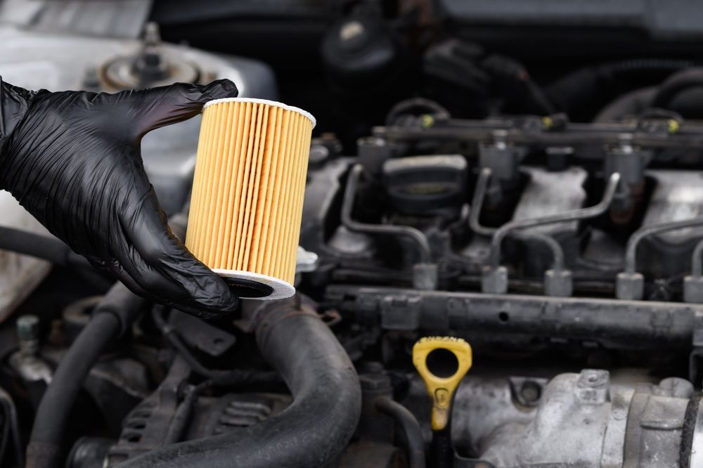 Una mano enguantada sostiene un filtro de aceite de motor nuevo, cilíndrico y amarillo, sobre el compartimento abierto del motor de un coche.