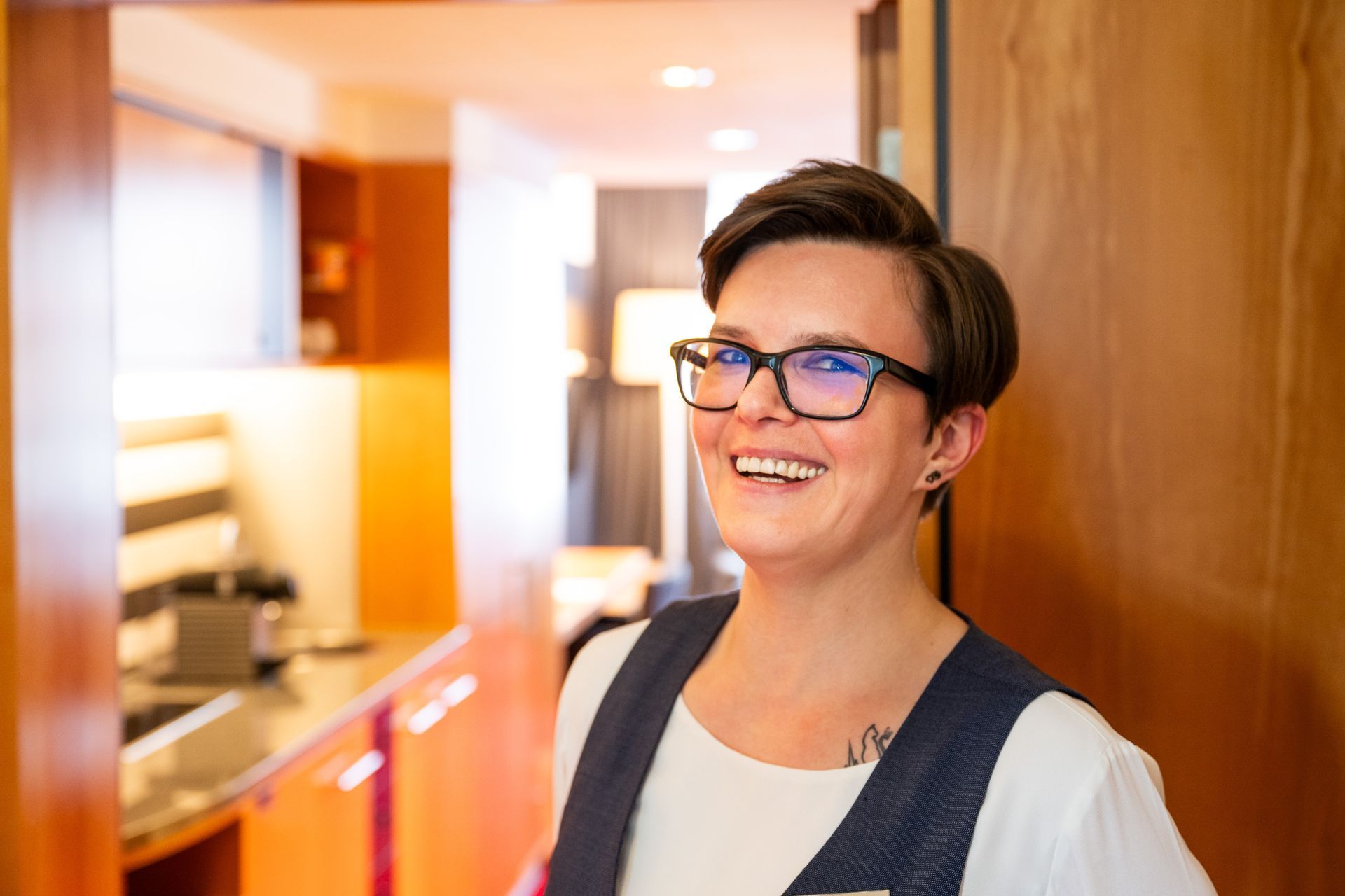 Eine Person mit kurzen dunklen Haaren und Brille steht lächelnd in einem Türrahmen. Sie trägt ein weißes Hemd und eine Weste.