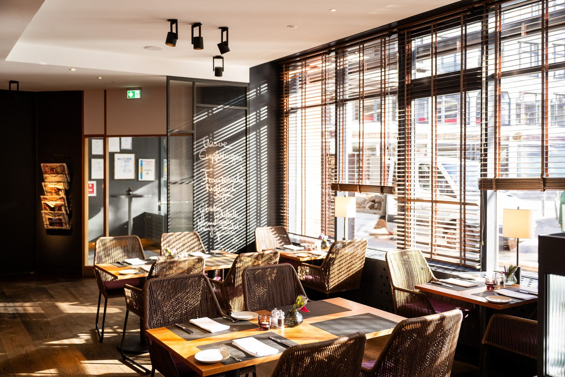 Restaurantinnenraum mit Tischen, Stühlen und großen Fenstern mit Jalousien.